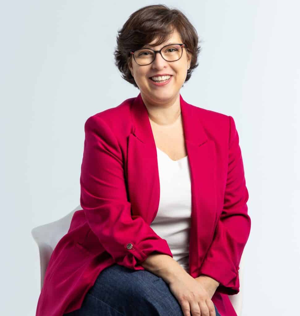 Una mujer de pelo castaño corto y gafas, vestida con una americana rosa brillante, un top blanco y vaqueros, está sentada sonriendo en una silla blanca sobre un fondo claro liso. Se encuentra en una consulta de psicología en Barcelona