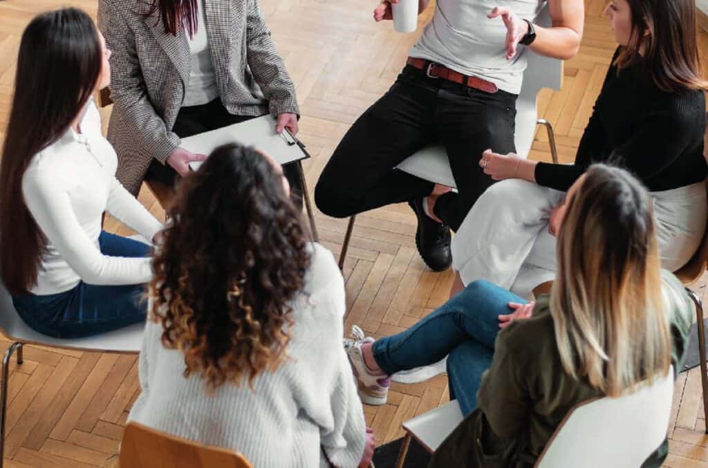 Seis personas sentadas en círculo sobre sillas, inmersas en una conversación o discusión, vistas desde arriba sobre un suelo de madera.