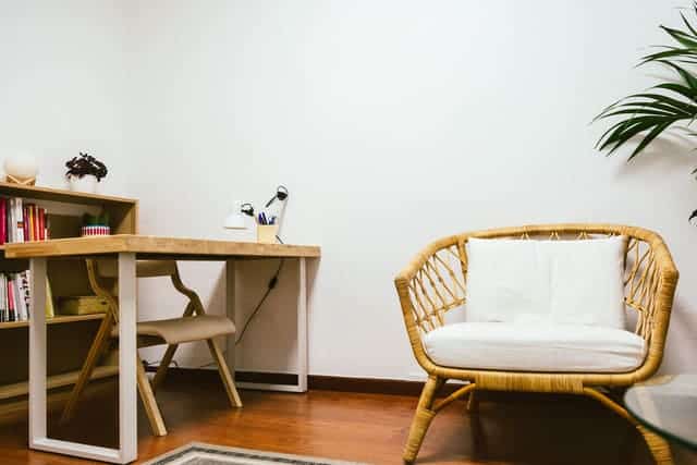 Una oficina doméstica minimalista con un escritorio y una silla de madera, un sillón de mimbre con cojines blancos, una estantería y una maceta en un rincón. En esta oficina se ofrece Apoyo psicológico en Barcelona
